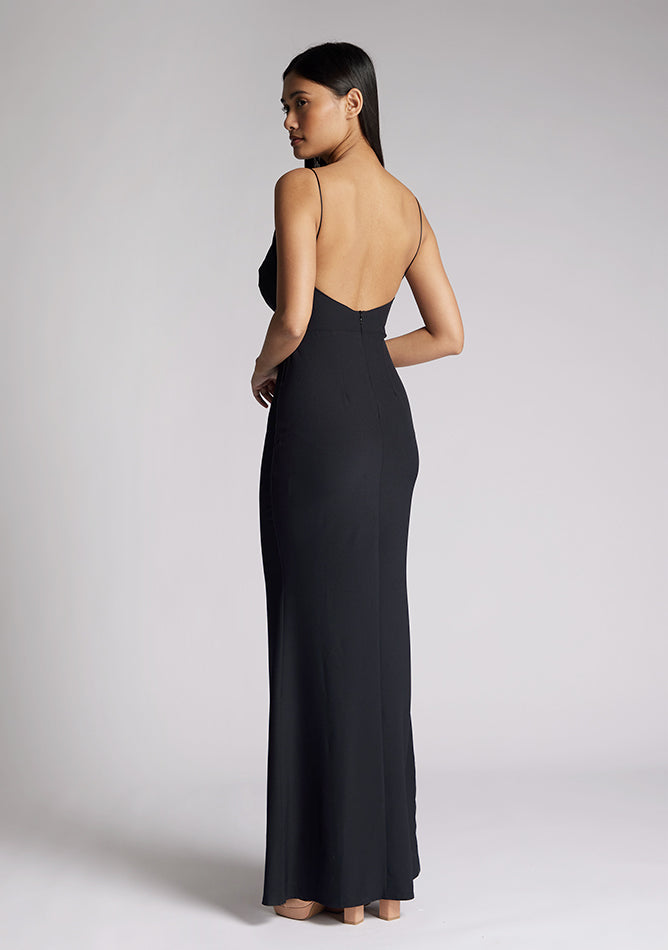 Back quarter image of a model wearing a black maxi dress with a v-neckline, thin straps with twist front detail at bust, low back and subtle flair at hem. The design featured is the Vesper Marlowe Black Maxi Dress