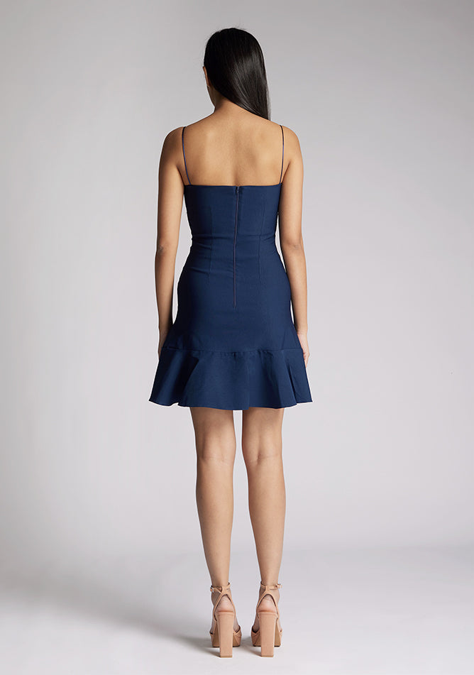 Back image of a model wearing a navy mini dress with a straight neckline, thin straps, a fitted bodice and flared ruffle hem with invisible centre back zip. The design featured is the Vesper Brenda Navy Mini Dress