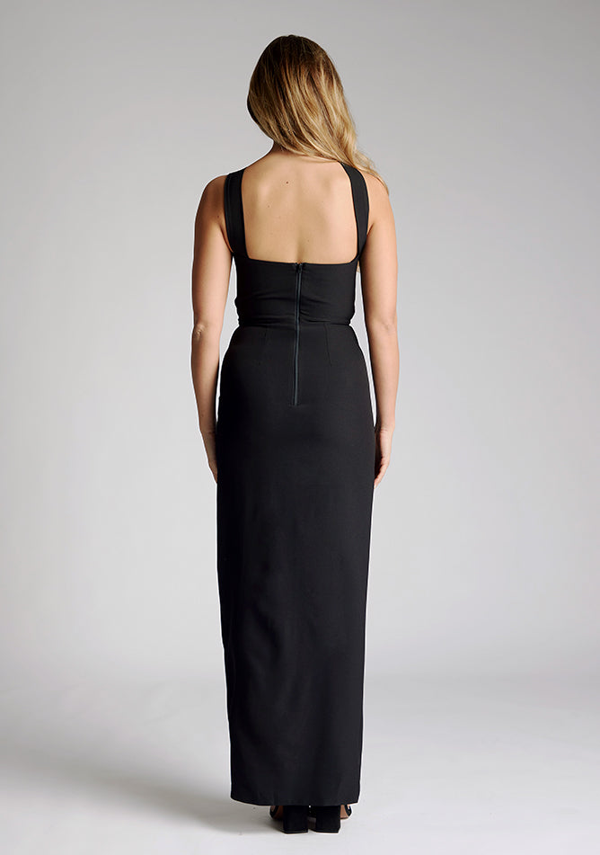 Back image of a model wearing a black maxi dress with a featuring front split and one shoulder strap with another going across the neck. The design featured is the Vesper Gizelle black maxi dress