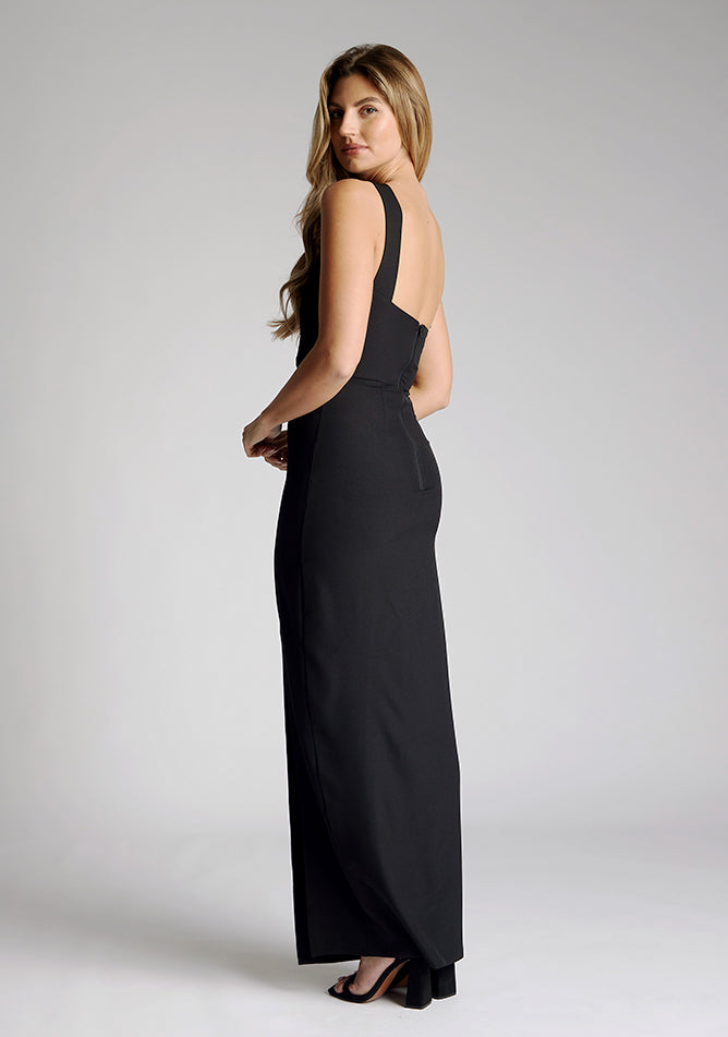 Back quarter image of a model wearing a black maxi dress with a featuring front split and one shoulder strap with another going across the neck. The design featured is the Vesper Gizelle black maxi dress