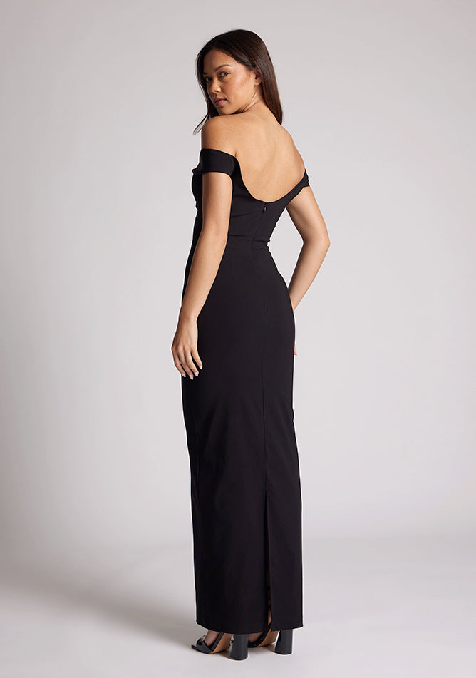 Back quarter image of a model wearing a black maxi dress, featuring off-the-shoulder short sleeves and a dipped back design. The dress feature is the Vesper Holland Black Maxi Bardot Dress.