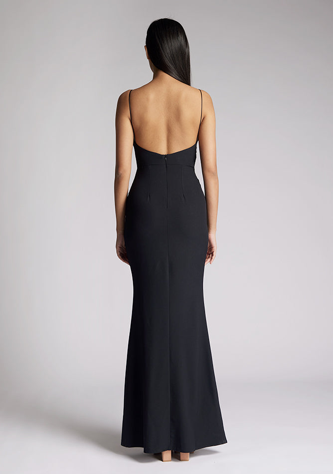 Back iimage of a model wearing a black maxi dress with a v-neckline, thin straps with twist front detail at bust, low back and subtle flair at hem. The design featured is the Vesper Marlowe Black Maxi Dress