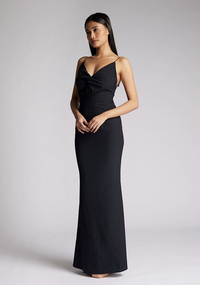 Front quarter image of a model wearing a black maxi dress with a v-neckline, thin straps with twist front detail at bust, low back and subtle flair at hem. The design featured is the Vesper Marlowe Black Maxi Dress