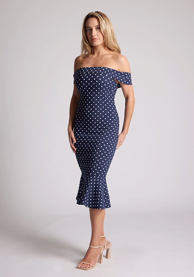 Front quarter image of the model wearing a navy polka bardot midaxi dress with a classy bardot neckline with arm bands and frill hem detail. The design featured is the Vesper Racquel Navy Polka Dot Bardot Midaxi Dress