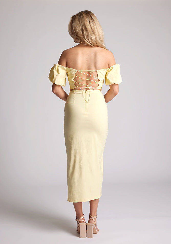 Back image of a blonde model wearing a yellow bardot midaxi dress with a subtle sweetheart neckline, statement puff sleeves, a front skirt split and lace-up detail at the back. The design featured is the Vesper River Yellow Bardot Midaxi Dress