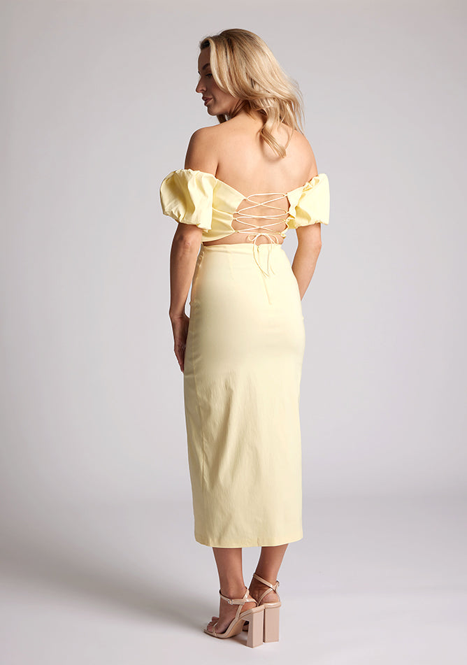 Back quarter image of a blonde model wearing a yellow bardot midaxi dress with a subtle sweetheart neckline, statement puff sleeves, a front skirt split and lace-up detail at the back. The design featured is the Vesper River Yellow Bardot Midaxi Dress