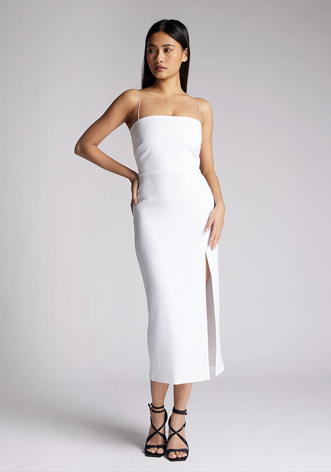 Front image of a model wearing a white midaxi dress with a minimalistic strap details, front skirt split and centre back zip. The design featured is the Vesper Tate White Midaxi Dress