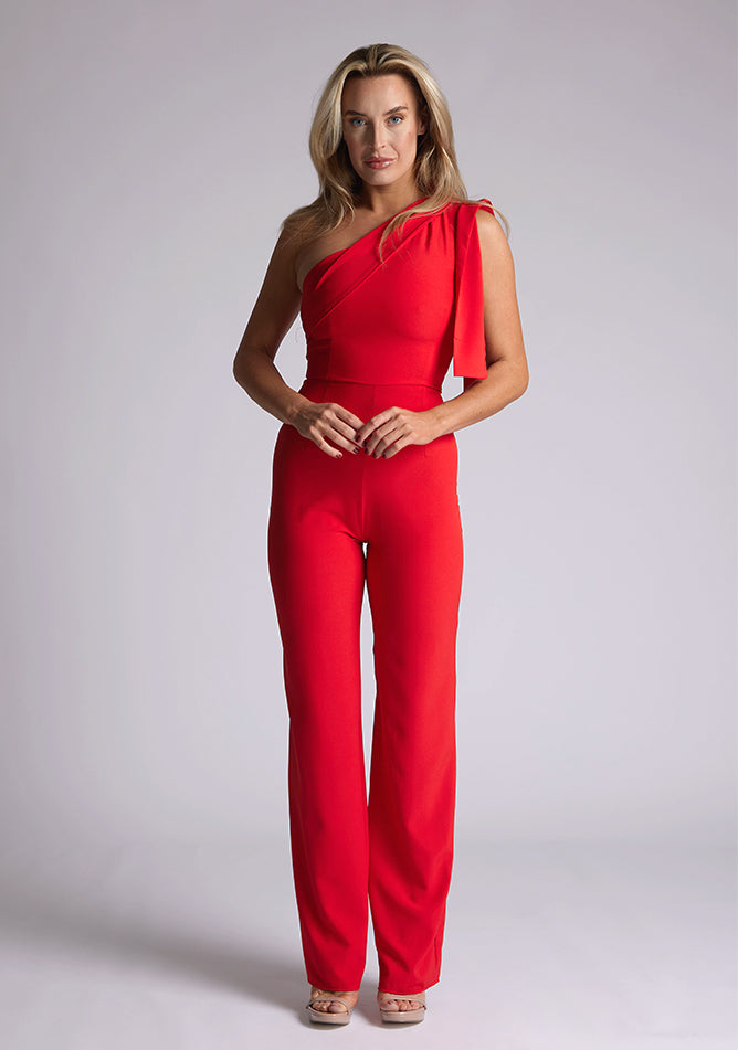 Front image of the model wearing a Red Jumpsuit with an asymmetric neckline and a bow on the shoulder. The design feature is the Vesper Gracie Red Jumpsuit.