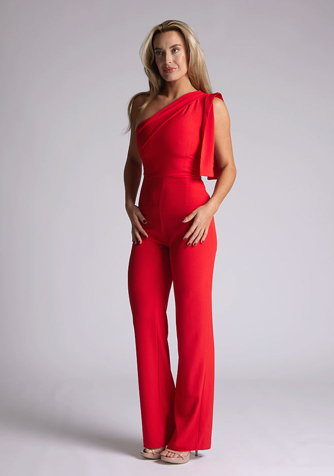 Front quarter image of the model wearing a Red Jumpsuit with an asymmetric neckline and a bow on the shoulder. The design feature is the Vesper Gracie Red Jumpsuit.