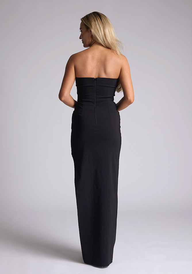 Back image of blonde model wearing a black maxi dress with a features a sweetheart neckline, centre back zip and a side skirt split. The design featured is the Vesper Larissa Black Bodycon Maxi Dress