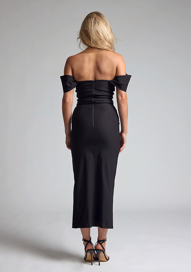 Back image of blonde model wearing a Black Midaxi Dress with a off-shoulder design, a flattering body-con sillhoutte, and a front skirt split,. The design features is the Vesper Marriot Black Bardot Midaxi Dress