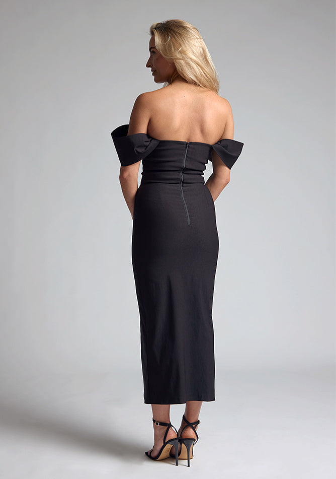 Back quarter image of blonde model wearing a Black Midaxi Dress with a off-shoulder design, a flattering body-con sillhoutte, and a front skirt split,. The design features is the Vesper Marriot Black Bardot Midaxi Dress