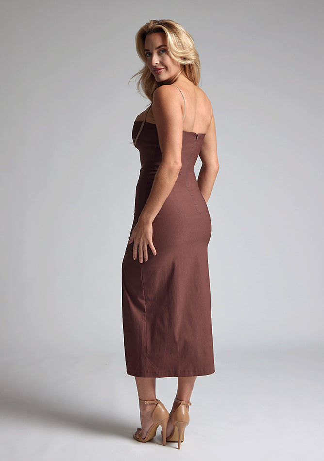 Back quarter image of a blonde model wearing a Chocolate Midaxi Dress with a daring plunge neckline, delicate thin straps, a flattering bodycon fit and a faux wrap skirt split adds a touch of sophistication to the overall design. The design features the Vesper Oaklynn Chocolate Midaxi Dress