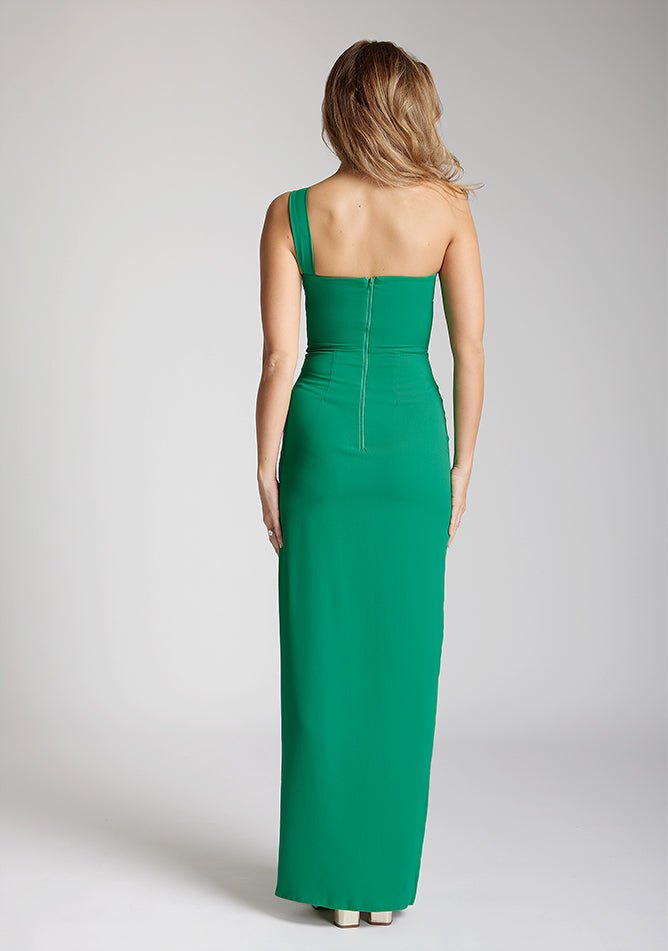 Back image of a blonde model wearing a emerald green maxidress with a asymmetric neckline, front skirt split, and beautiful emerald green hue. The design featured is the Vesper Olivette Emerald Green Maxi Dress