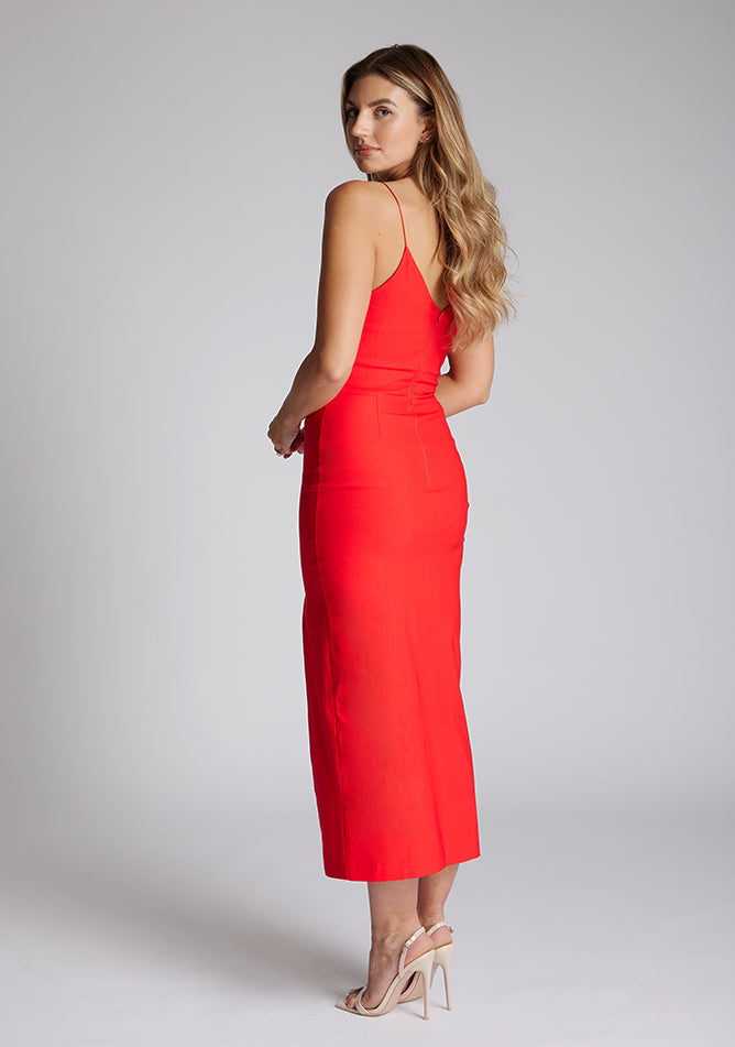 Back quarter image of blonde model wearing a red midaxi dress with a plunge neckline, delicate thin straps for a feminine touch, and front split that adds a modern edge. The design featured is the Vesper Palmer Red Midaxi Dress