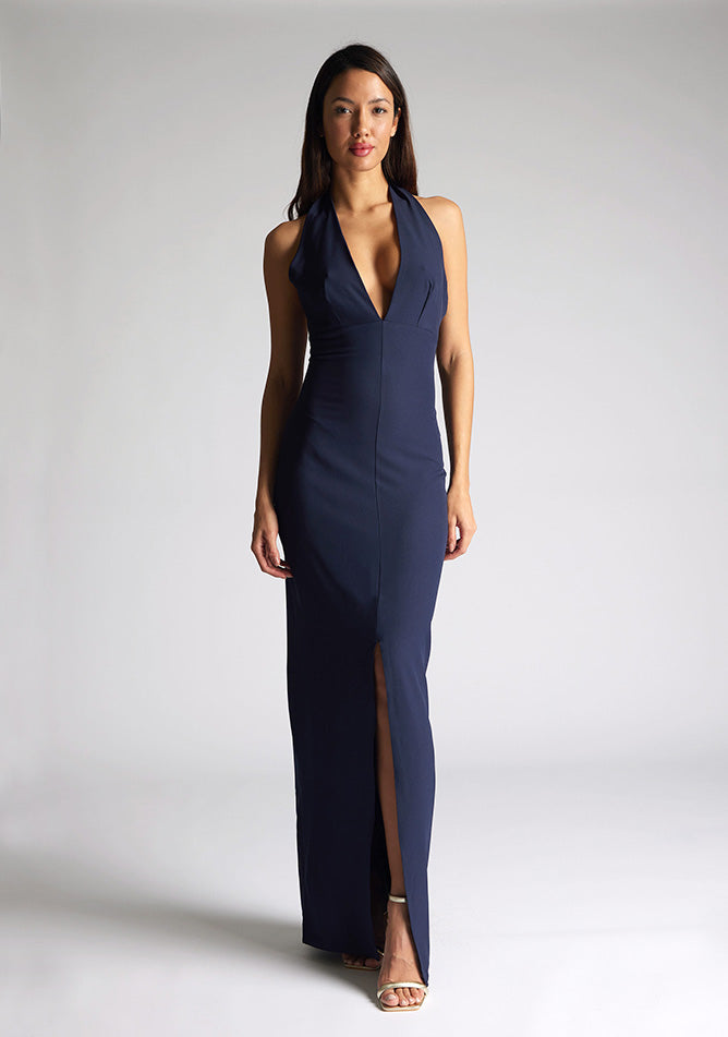 Front image of a blonde model wearing a navy maxidress with a v-plunging neckline, halter design and a front skirt split. The design featured is the Vesper Sedona Navy Maxi Dress