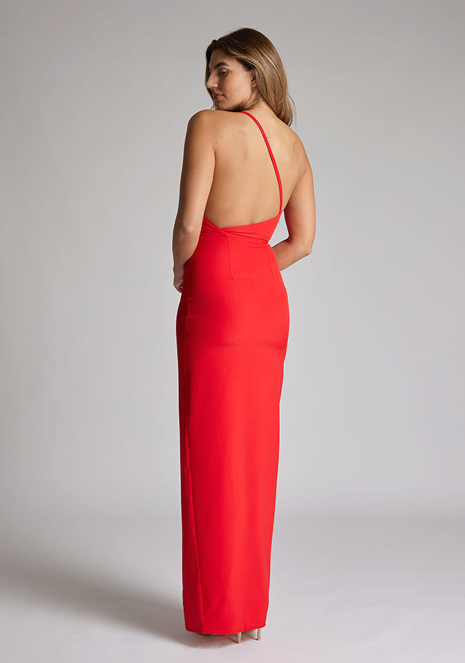Back quarter image of a model wearing a red maxi dress with a asymmetric neckline, one shoulder strap, front skirt split and open back with asymmetric strap across back. The design featured is Vesper Viva Red Maxi Dress