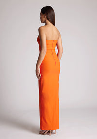 Back quarter image of a model wearing a orange strapless maxi dress with a front split and centre back zip. The design featured is the Vesper Alaya strapless maxi dress