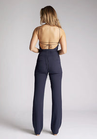 Back image of a model wearing a backless navy halter neck jumpsuit, featuring a key hole cut out and straps which cross at the back. The design featured is the Vesper Amanda navy jumpsuit