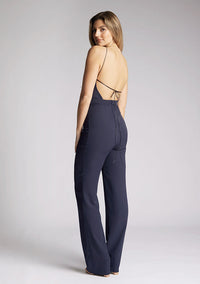 Back quarter image of a model wearing a backless navy halter neck jumpsuit, featuring a key hole cut out and straps which cross at the back. The design featured is the Vesper Amanda navy jumpsuit
