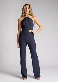 Front image of a model wearing a backless navy halter neck jumpsuit, featuring a key hole cut out and straps which cross at the back. The design featured is the Vesper Amanda navy jumpsuit