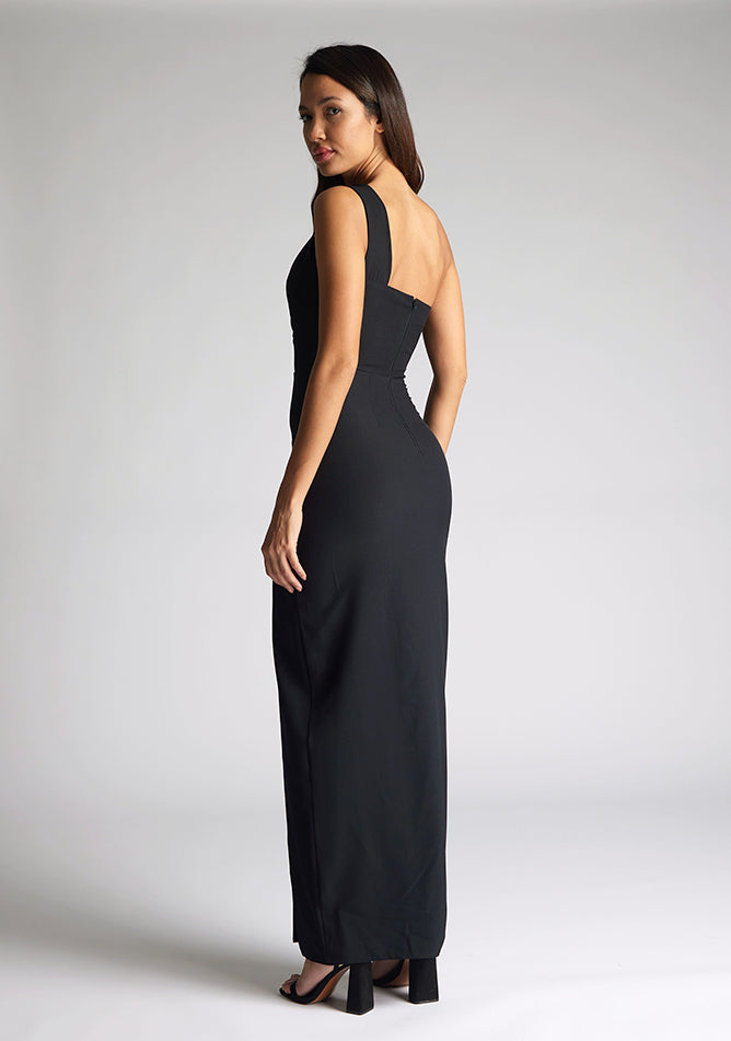 Back quarter image of the model wearing a Black Maxi Dress with a one-shoulder design with a front cut-out, and a front skirt split, a design features Vesper Ellis Black Maxi Dress