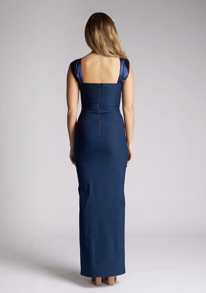 Back image of a model wearing a navy maxi dress, featuring a body con design and a satin panel at the top which twists up into straps. The dress featured is the Vesper Fame navy maxi dress
