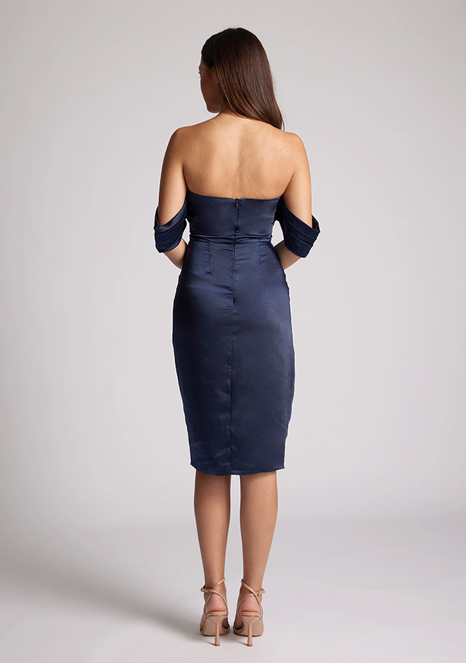 Back image of a model wearing a navy satin midi dress, featuring a pleated panel across the front and two arm bands. The dress featured is the Vesper Fleur navy midi dress