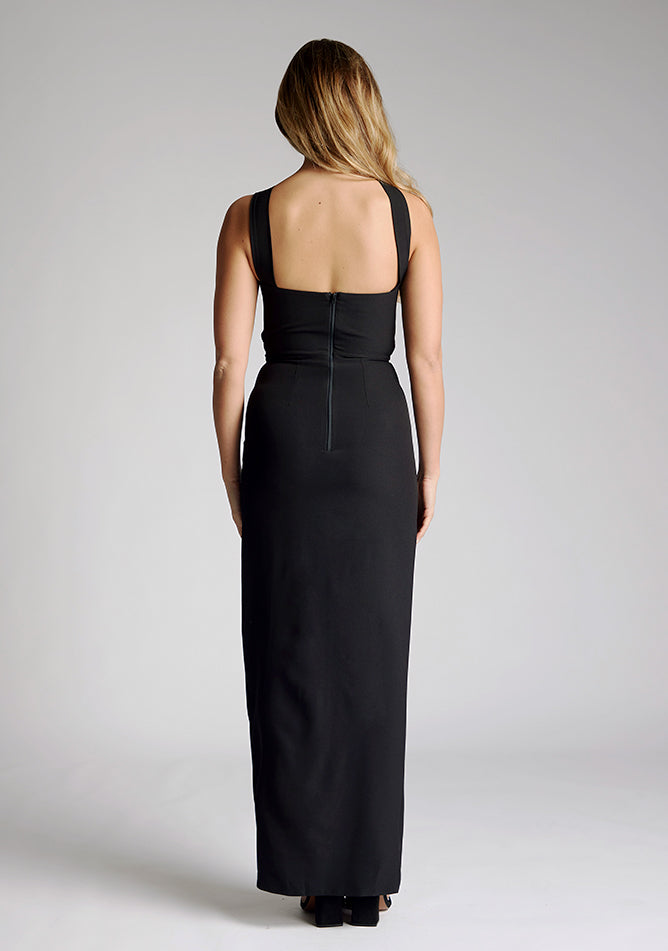 Back image of a model wearing a black maxi dress, featuring a front split and one shoulder strap with another going across the neck. The dress featured is the Vesper Gizelle black maxi dress