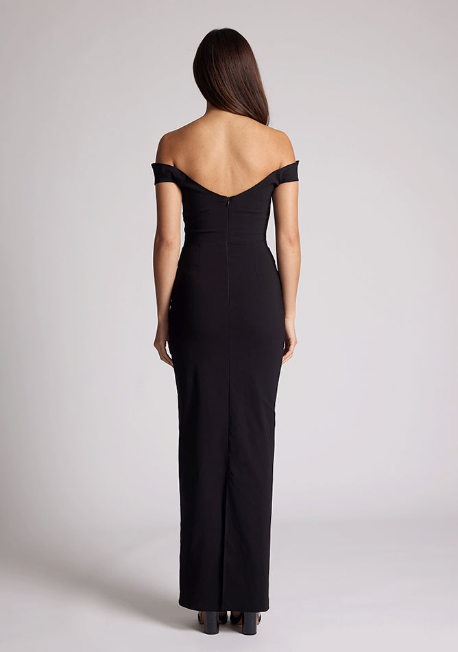 Back image of a model wearing a black maxi dress, featuring off the shoulder short sleeves and a dipped back design. The dress featured is the Vesper Holland black maxi dress