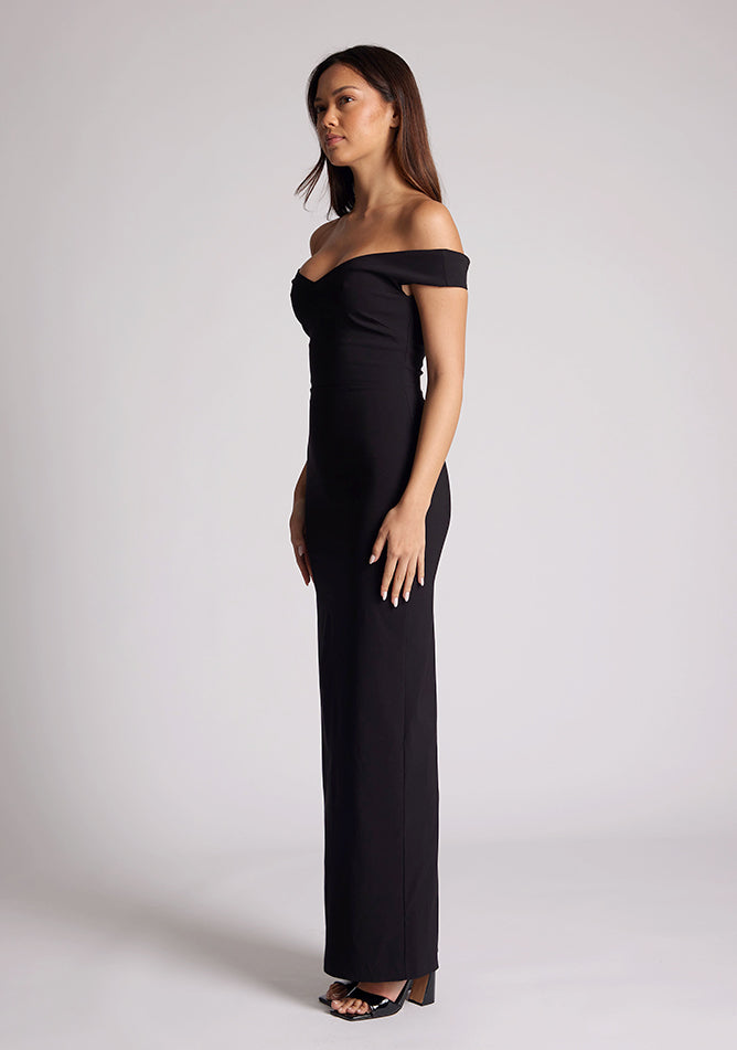 Front quarter image of a model wearing a black maxi dress, featuring off the shoulder short sleeves and a dipped back design. The dress featured is the Vesper Holland black maxi dress