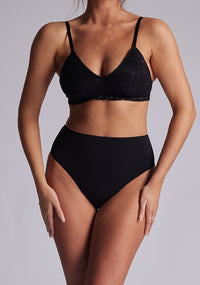 A front image of a model wearing a a black bra with a lace overlay and lace back. The bra featured is the Midnight Lace Overlay Bra and is worn with the Opal Lace High Waist Bottoms