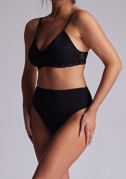 A side image of a model wearing a a black bra with a lace overlay and lace back. The bra featured is the Midnight Lace Overlay Bra and is worn with the Opal Lace High Waist Bottoms