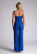Back image of a model wearing a royal blue jumpsuit, featuring a sweetheart neckline and tie up straps. The jumpsuit featured is the Vesper Nicole royal blue tie up jumpsuit
