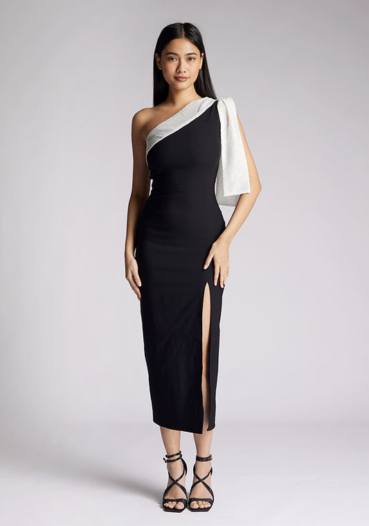 Front image of a model wearing a monochrome midaxi dress, featuring an asymmetric neckline with bow detail at shoulder in ivory sequin, front skirt split and centre back invisible zip on the black dress. The dress featured is the Vesper Odette Monochrome Sequin Midaxi Dress