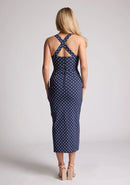 Back image of a model wearing a navy polka dot midaxi dress, featuring a sweetheart neckline and straps that cross at the back. The dress featured is the Vesper Poppy navy polka dot sweetheart midaxi dress