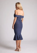 Back quarter image of a model wearing a navy polka dot dress, featuring a bardot neckline and frill hem. The dress featured is the Vesper Racquel navy polka dot bardot midaxi dress