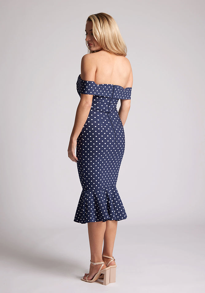 Back quarter image of a model wearing a navy polka dot dress, featuring a bardot neckline and frill hem. The dress featured is the Vesper Racquel navy polka dot bardot midaxi dress
