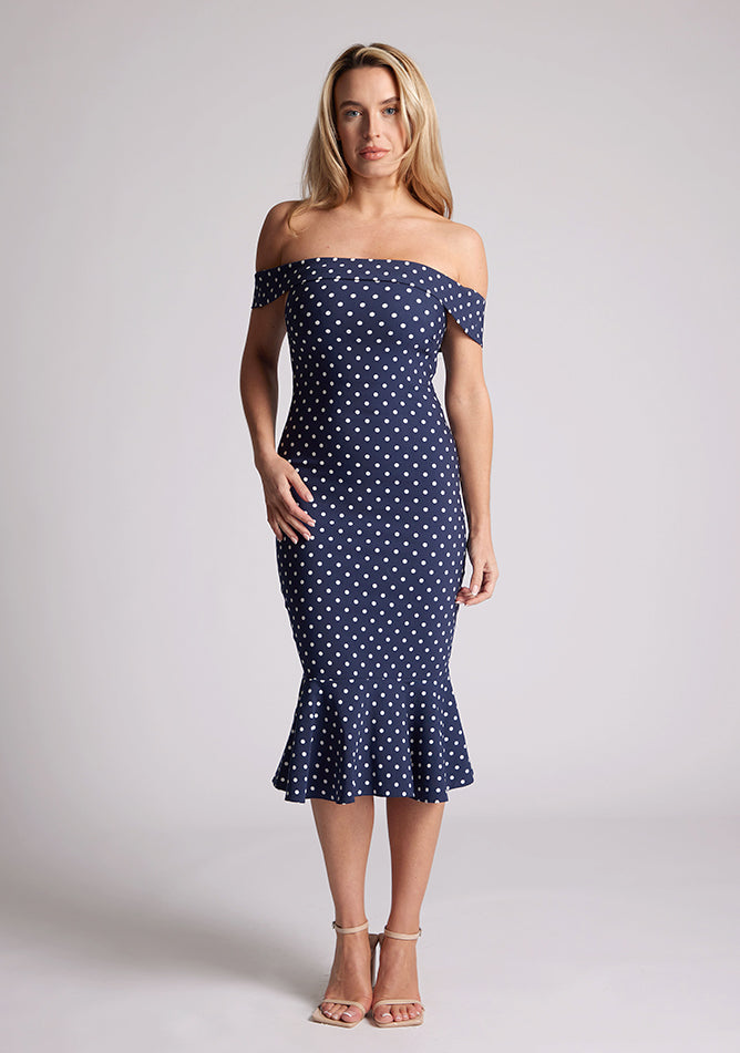 Front image of a model wearing a navy polka dot dress, featuring a bardot neckline and frill hem. The dress featured is the Vesper Racquel navy polka dot bardot midaxi dress