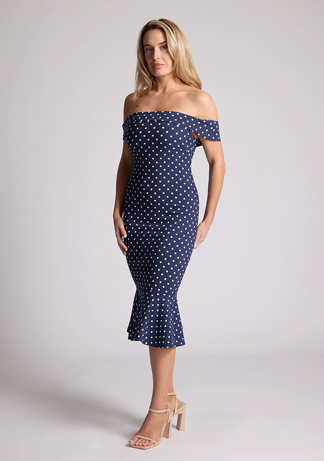 Front quarter image of a model wearing a navy polka dot dress, featuring a bardot neckline and frill hem. The dress featured is the Vesper Racquel navy polka dot bardot midaxi dress
