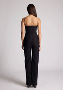 Back image of a model wearing a black jumpsuit, featuring a sweetheart neckline and a wide leg design. The jumpsuit featured is the Vesper Rogan black jumpsuit