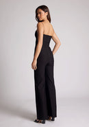 Back quarter image of a model wearing a black jumpsuit, featuring a sweetheart neckline and a wide leg design. The jumpsuit featured is the Vesper Rogan black jumpsuit