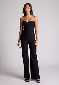 Front image of a model wearing a black jumpsuit, featuring a sweetheart neckline and a wide leg design. The jumpsuit featured is the Vesper Rogan black jumpsuit
