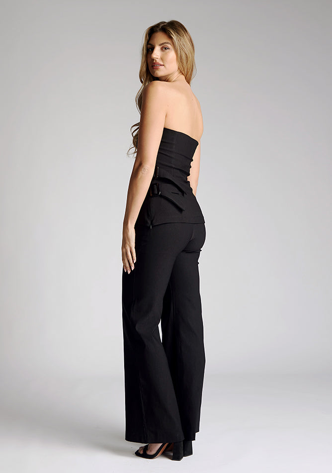 Back quarter image of a model wearing a strapless black top, featuring an asymmetric hem and buckle detailing. The top featured is the Vesper Sharon black top worn with the Vesper June black wide leg trousers