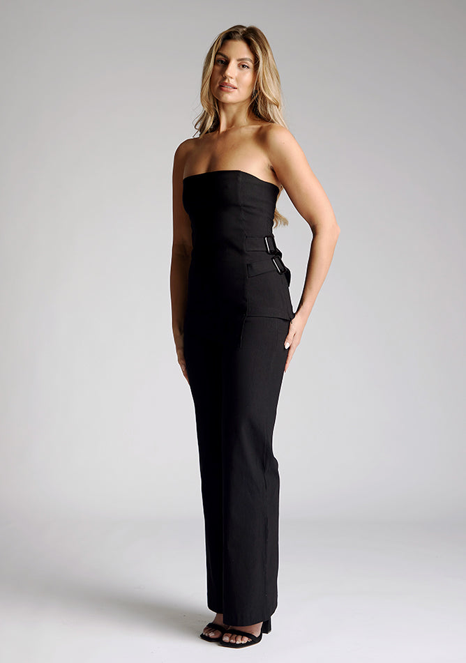 Front quarter image of a model wearing a strapless black top, featuring an asymmetric hem and buckle detailing. The top featured is the Vesper Sharon black top worn with the Vesper June black wide leg trousers