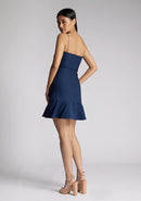 Back quarter image of a model wearing a navy mini dress with a straight neckline, thin straps, a fitted bodice and flared ruffle hem with invisible centre back zip. The design featured is the Vesper Brenda Navy Mini Dress