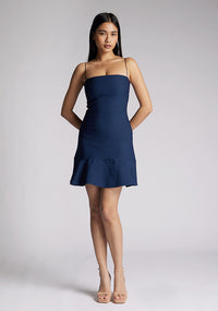 Front image of a model wearing a navy mini dress with a straight neckline, thin straps, a fitted bodice and flared ruffle hem with invisible centre back zip. The design featured is the Vesper Brenda Navy Mini Dress