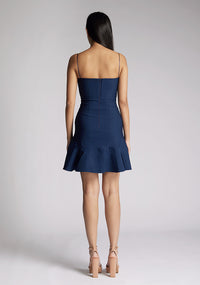 Back image of a model wearing a navy mini dress, featuring a straight neckline, thin straps, a fitted bodice and flared ruffle hem with invisible centre back zip. The dress featured is the Vesper Brenda Navy Mini Dress.