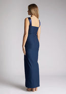 Back quarter image of a model wearing navy maxi dress with a chic neckline, sleeveless silhouette, figure-hugging body con design and satin panel at the top which twists up into straps. The design featured is the Vesper Fame Navy Maxi Dress
