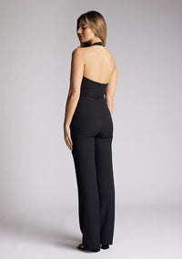 Back quarter image of a model wearing a black jumpsuit, featuring deep v-neckline with halter neck style straps, a wide leg and gold buttons at beck of neck. The style featured is Vesper Hazel Black Halter Neck Jumpsuit